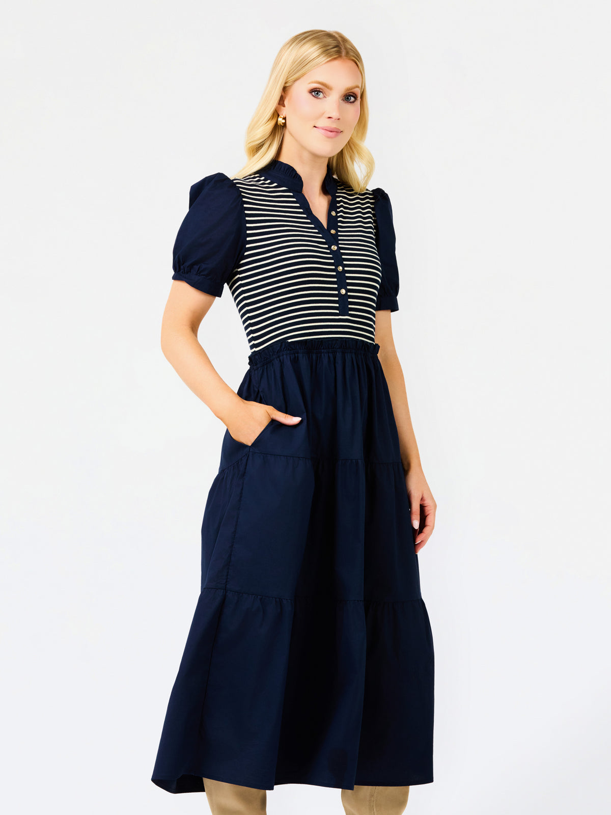 A woman modeling a navy blue midi dress with a navy and white striped bodice, short puff sleeves, and a tiered skirt, set against a plain white studio background.
