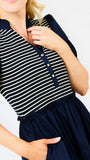 A close-up of a woman wearing a navy blue and white horizontally striped short-sleeve top with a buttoned henley-style neckline and a navy skirt, against a plain white background.