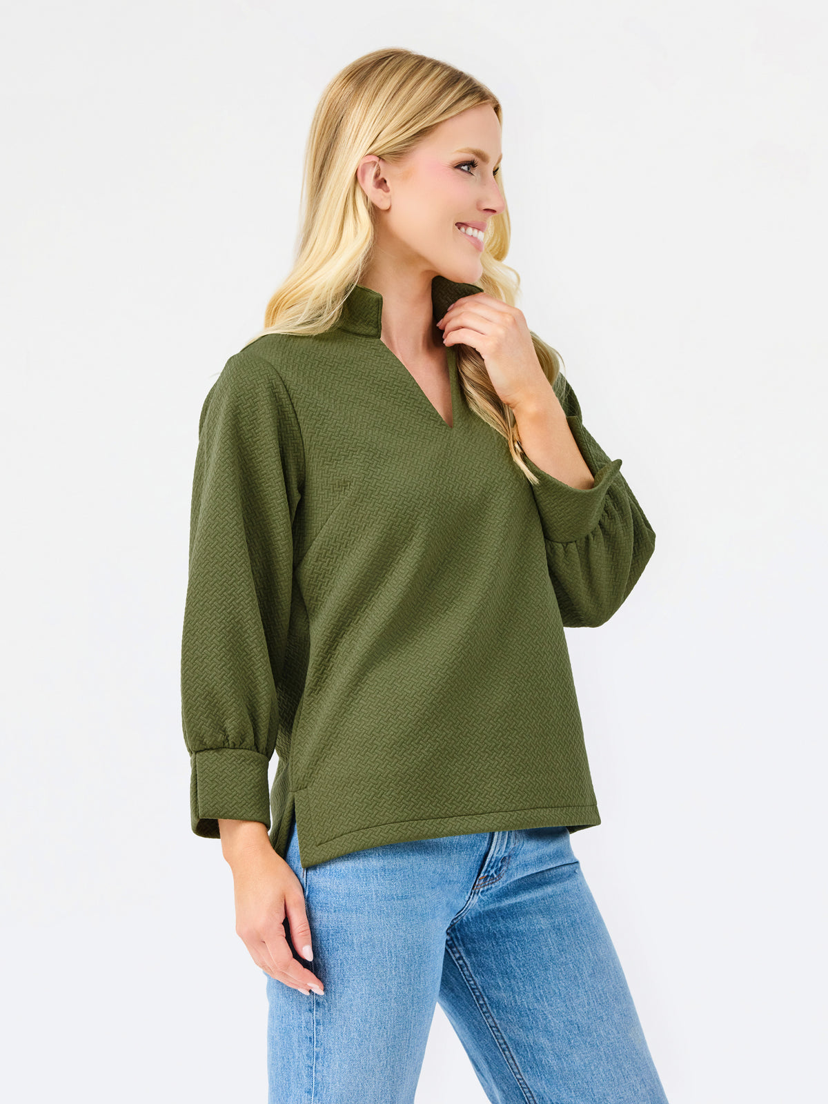 A woman wearing an olive green textured long-sleeve top with a split V-neck and blue jeans, standing against a plain white studio background.