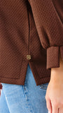 Close-up of a person wearing a brown textured jacket with a small gold button detail, paired with blue jeans.