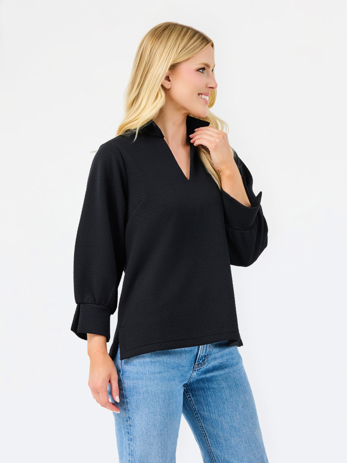A blonde woman wearing a black long-sleeve V-neck blouse with rolled cuffs and light blue jeans, standing against a plain white studio background.