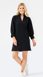 A woman modeling a black long-sleeve mini dress with a V-neckline and cuffed sleeves, paired with white sneakers, against a plain white studio background.