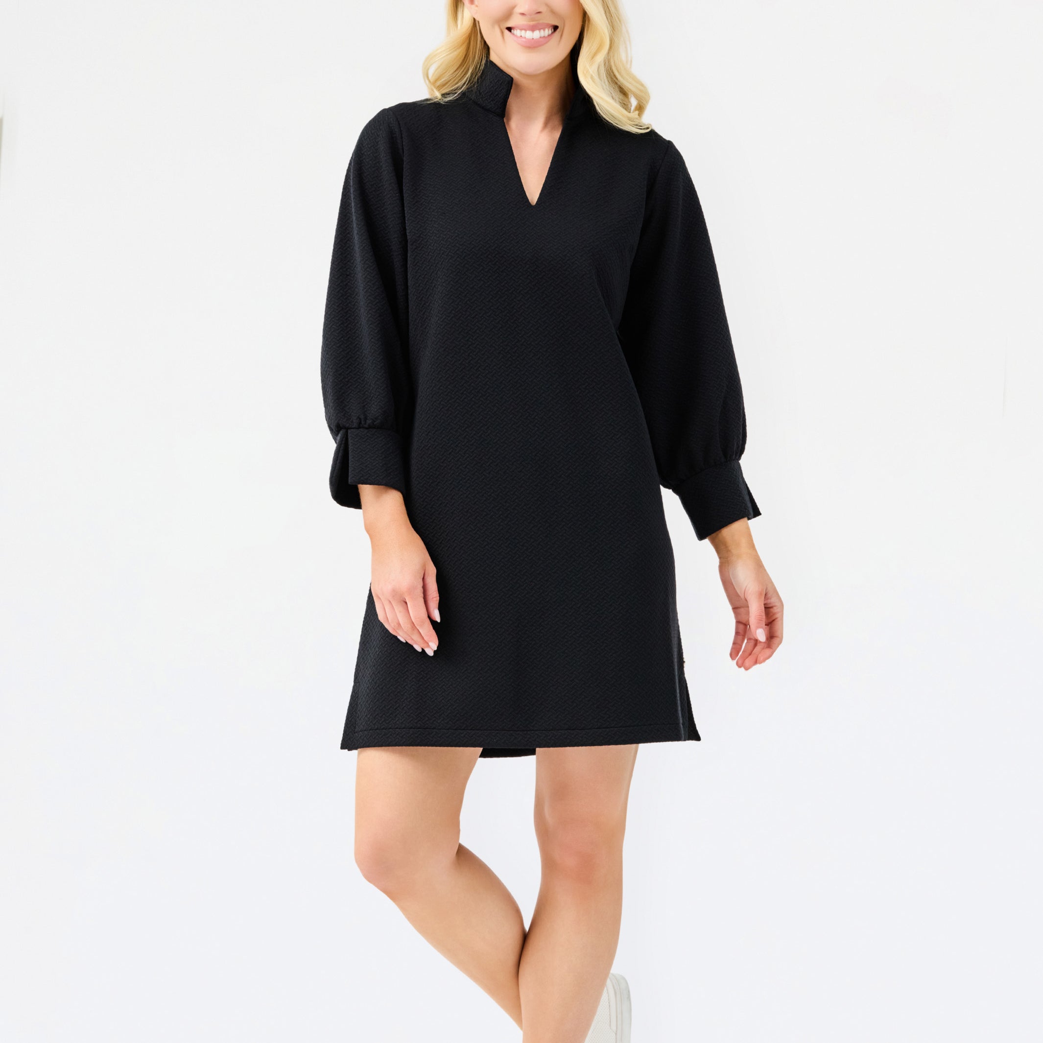A woman modeling a black long-sleeve mini dress with a V-neckline and cuffed sleeves, paired with white sneakers, against a plain white studio background.