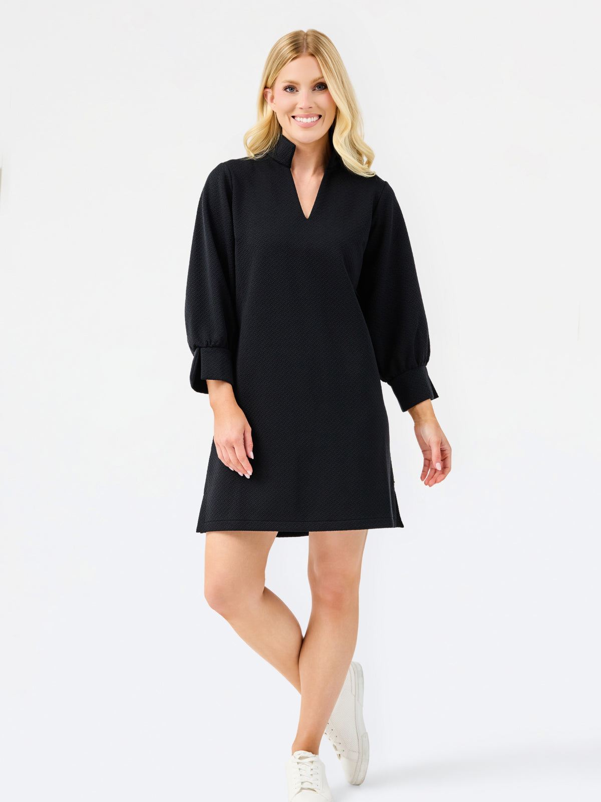 A woman modeling a black long-sleeve mini dress with a V-neckline and cuffed sleeves, paired with white sneakers, against a plain white studio background.