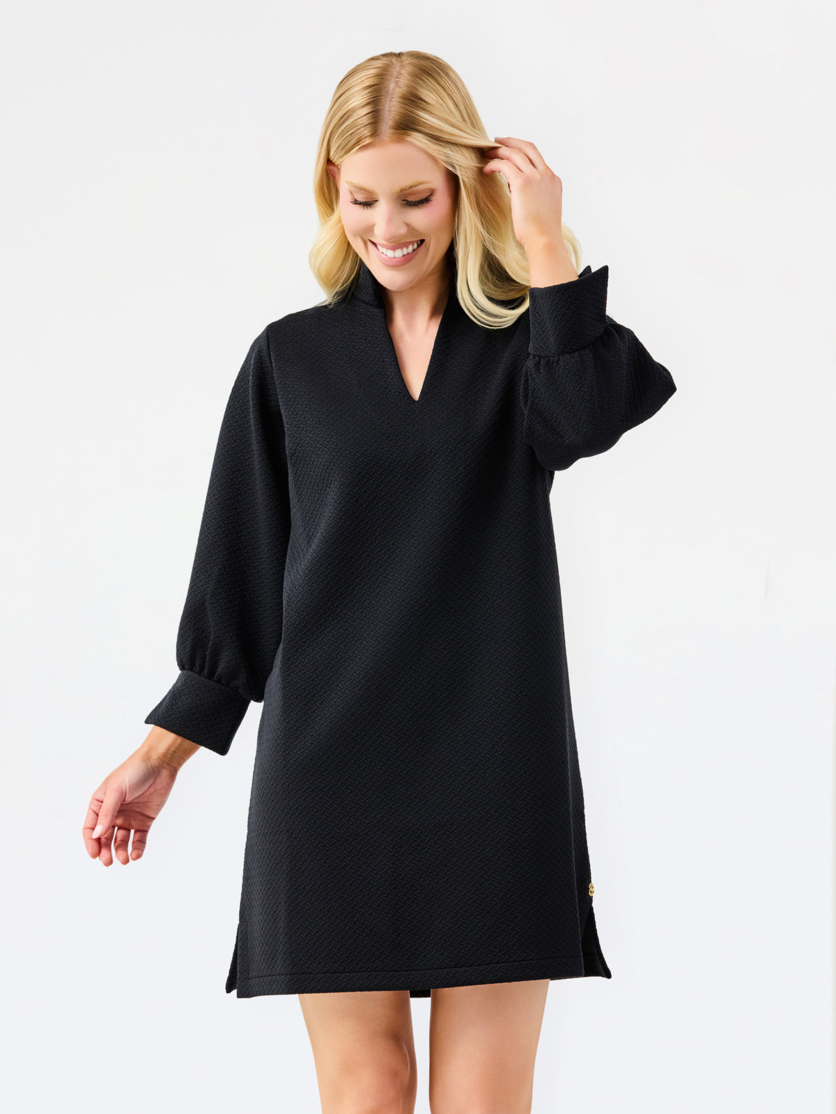 A blonde woman modeling a black long-sleeve mini dress with a V-neck and gathered cuffs, photographed against a clean white studio background.