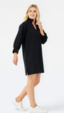 A woman modeling a black long-sleeve mini dress with a mock neck and side slit, paired with white sneakers, against a plain white studio background.