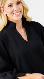 A smiling blonde woman wearing a black textured waffle-knit top with a mock neck and split V-neckline, photographed against a clean white background.