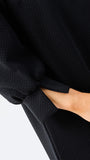 Close-up of a person wearing a black textured long-sleeve dress, with the sleeve cuff gathered and a hand inserted into a pocket, against a plain light background.