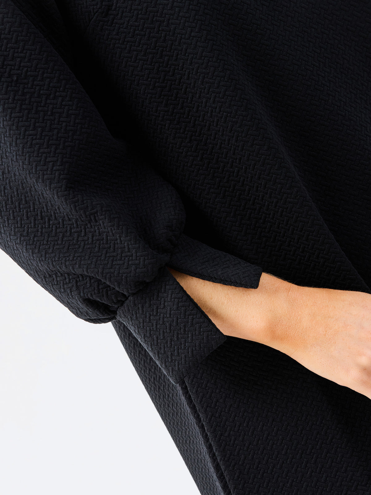 Close-up of a person wearing a black textured long-sleeve dress, with the sleeve cuff gathered and a hand inserted into a pocket, against a plain light background.