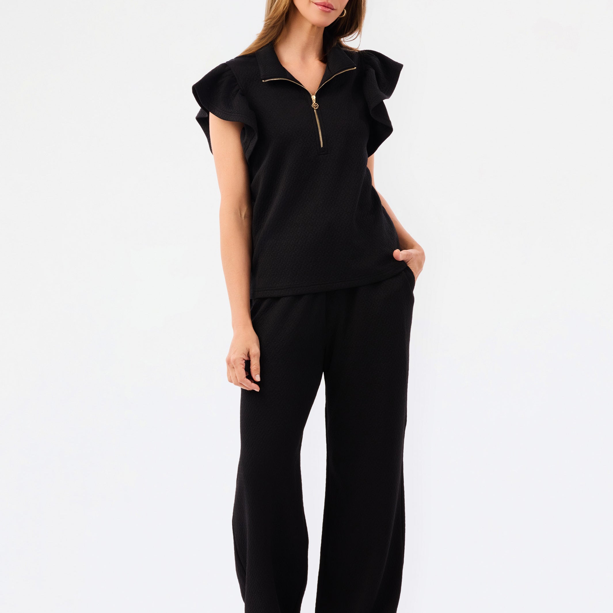 A woman modeling a black two-piece outfit consisting of a short-sleeve top with ruffled shoulders and a gold zipper, paired with matching wide-leg pants, against a plain white studio background.