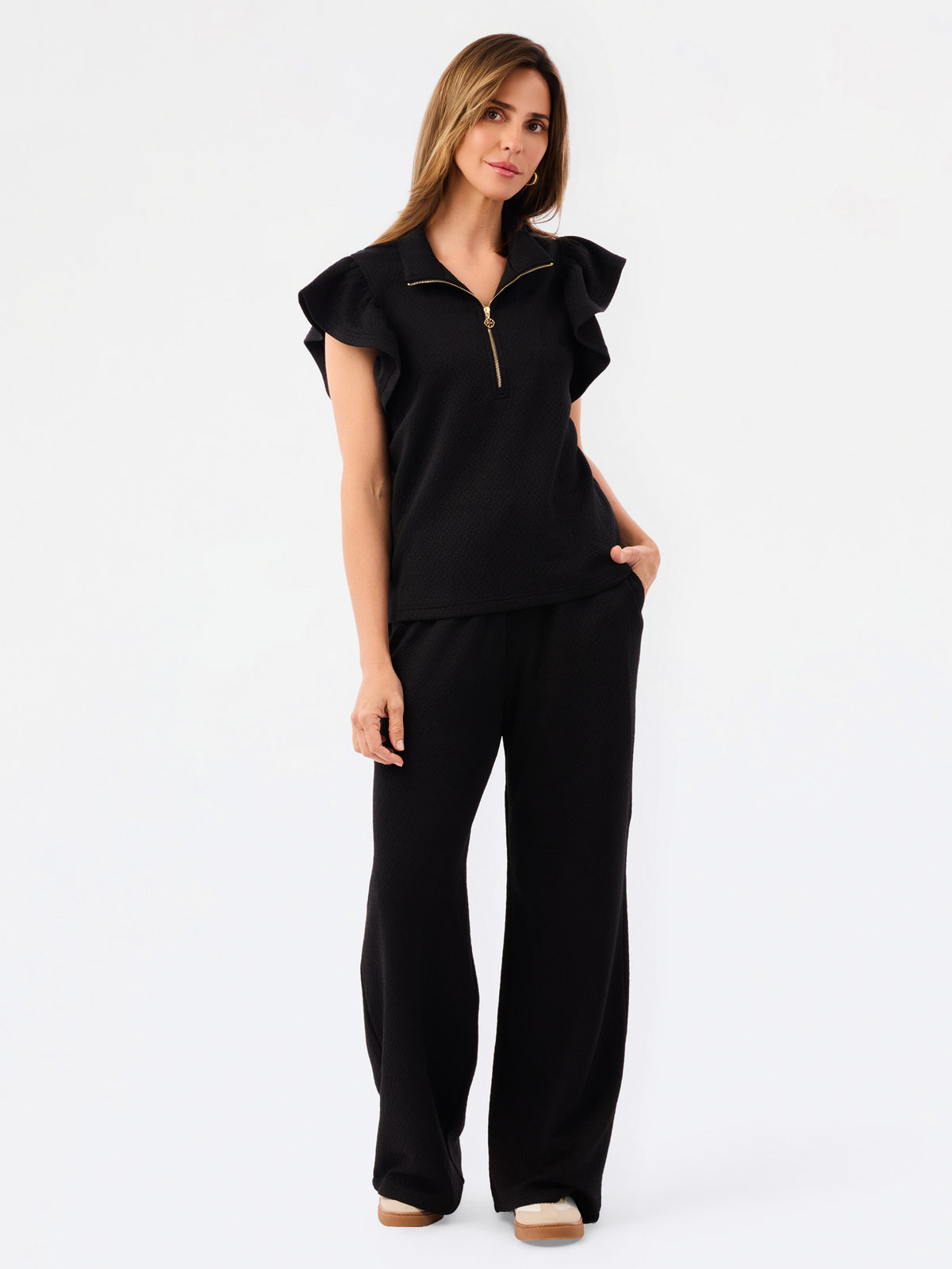 A woman modeling a black two-piece outfit consisting of a short-sleeve top with ruffled shoulders and a gold zipper, paired with matching wide-leg pants, against a plain white studio background.