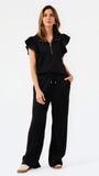 A woman modeling a black two-piece outfit consisting of a short-sleeve top with ruffled shoulders and a gold zipper, paired with matching wide-leg pants, against a plain white studio background.