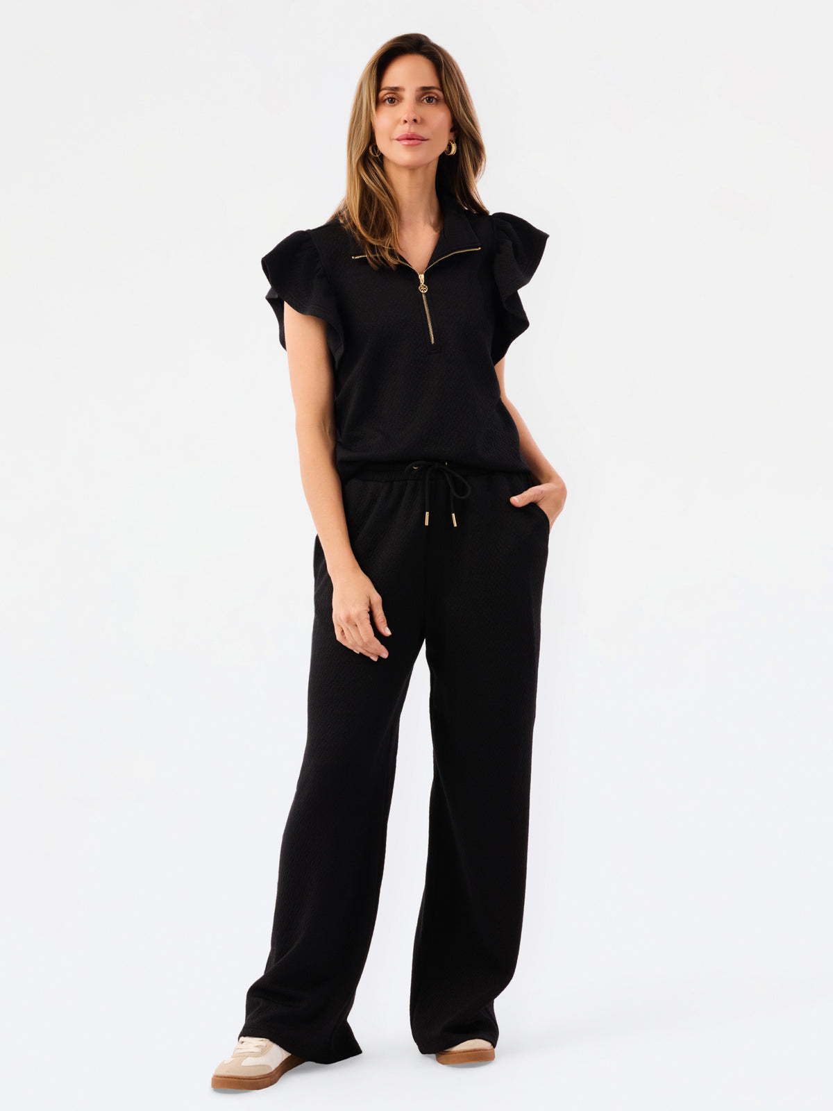 A woman modeling a black two-piece outfit consisting of a short-sleeve top with ruffled shoulders and a gold zipper, paired with matching wide-leg pants, against a plain white studio background.
