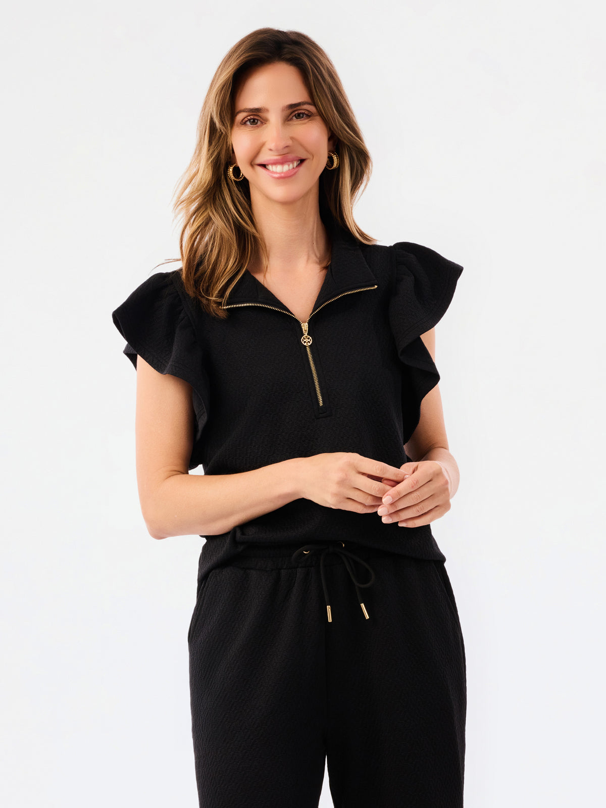 A woman modeling a black two-piece outfit consisting of a short-sleeve top with ruffled shoulders and a gold zipper, paired with matching wide-leg pants, against a plain white studio background.