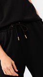 A woman modeling a black two-piece outfit consisting of a short-sleeve top with ruffled shoulders and a gold zipper, paired with matching wide-leg pants, against a plain white studio background.