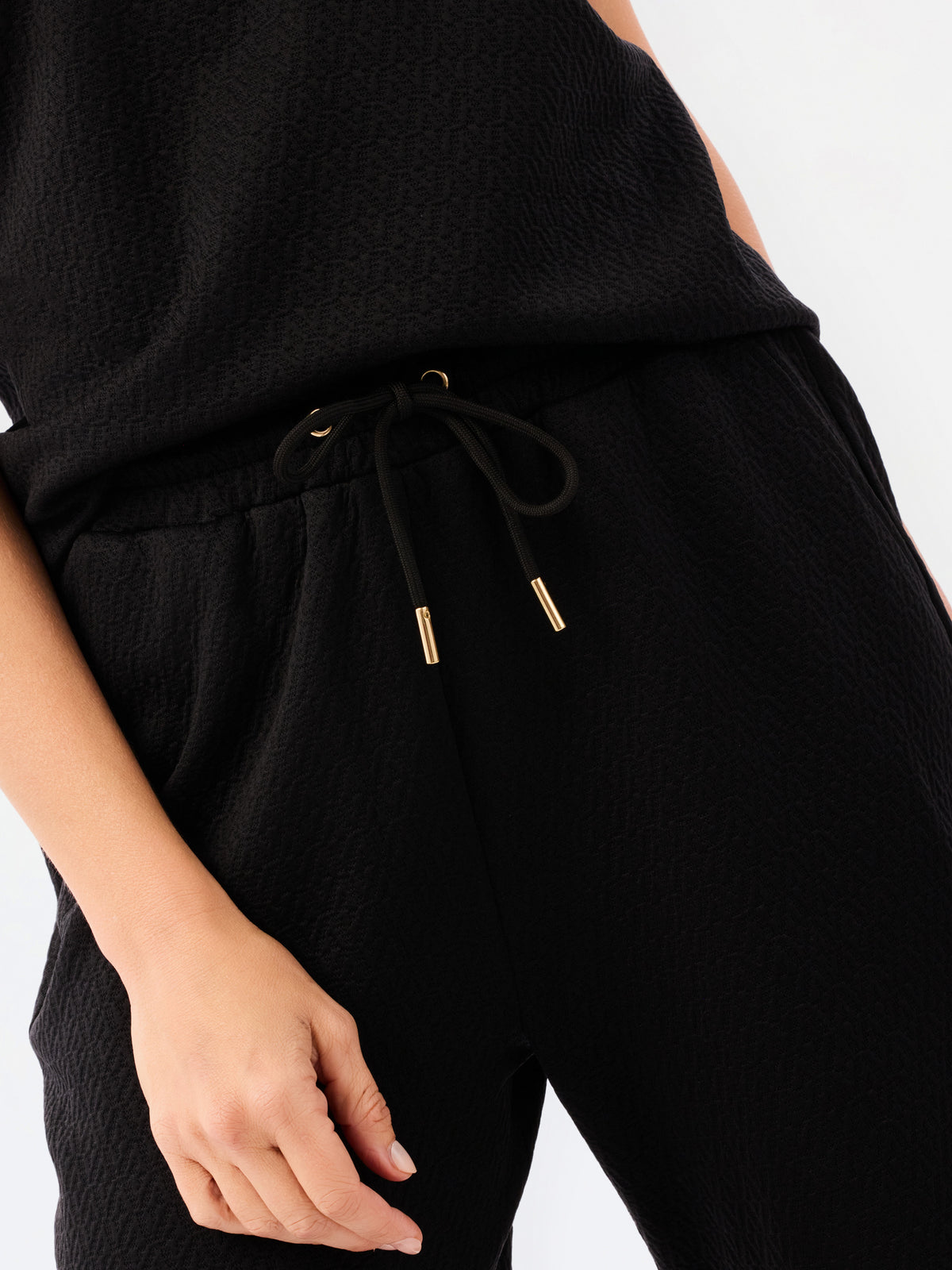 A woman modeling a black two-piece outfit consisting of a short-sleeve top with ruffled shoulders and a gold zipper, paired with matching wide-leg pants, against a plain white studio background.