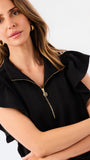 A woman modeling a black two-piece outfit consisting of a short-sleeve top with ruffled shoulders and a gold zipper, paired with matching wide-leg pants, against a plain white studio background.