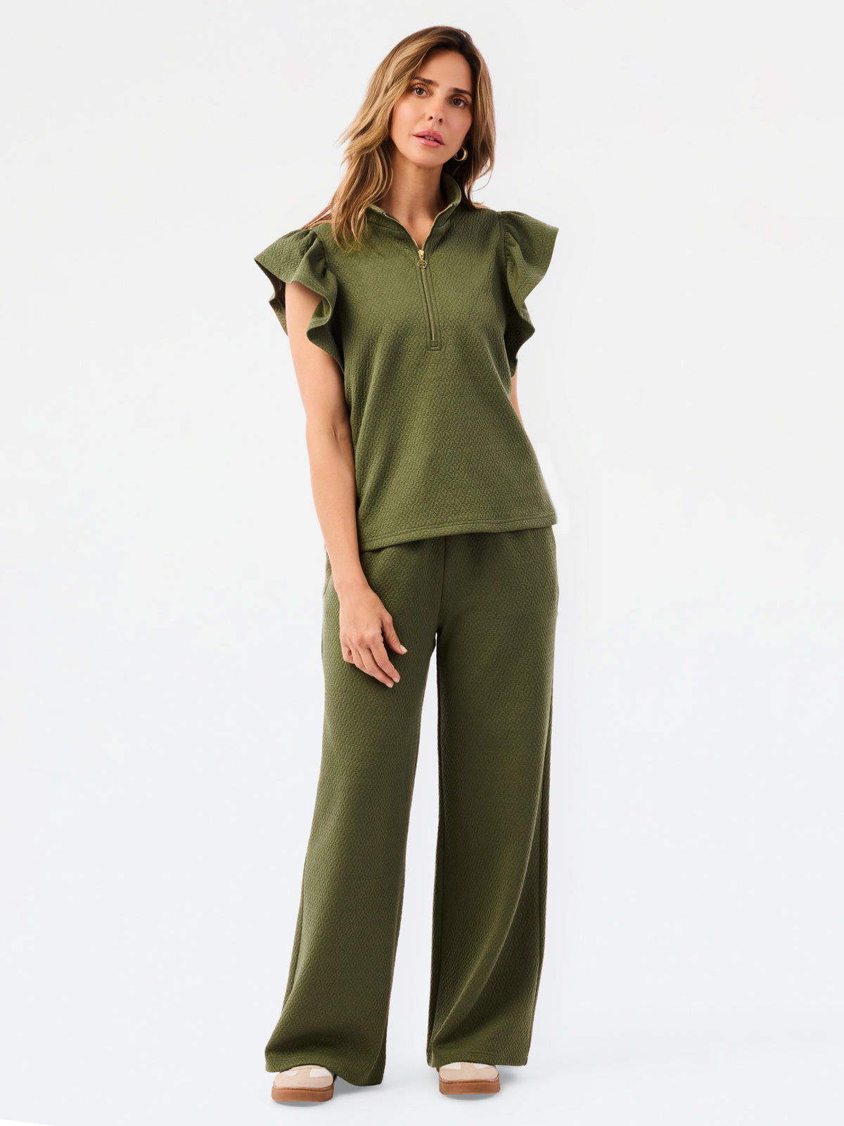 A woman modeling a matching olive green two-piece set consisting of a ruffled short-sleeve quarter-zip top and wide-leg pants, photographed against a clean white studio 