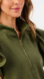 A woman modeling a matching olive green two-piece set consisting of a ruffled short-sleeve quarter-zip top and wide-leg pants, photographed against a clean white studio 