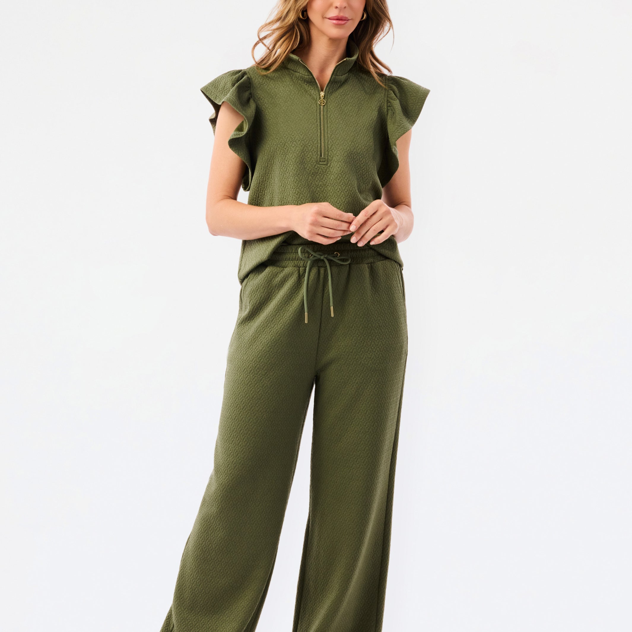 A woman modeling a matching olive green two-piece set consisting of a ruffled short-sleeve quarter-zip top and wide-leg pants, photographed against a clean white studio 