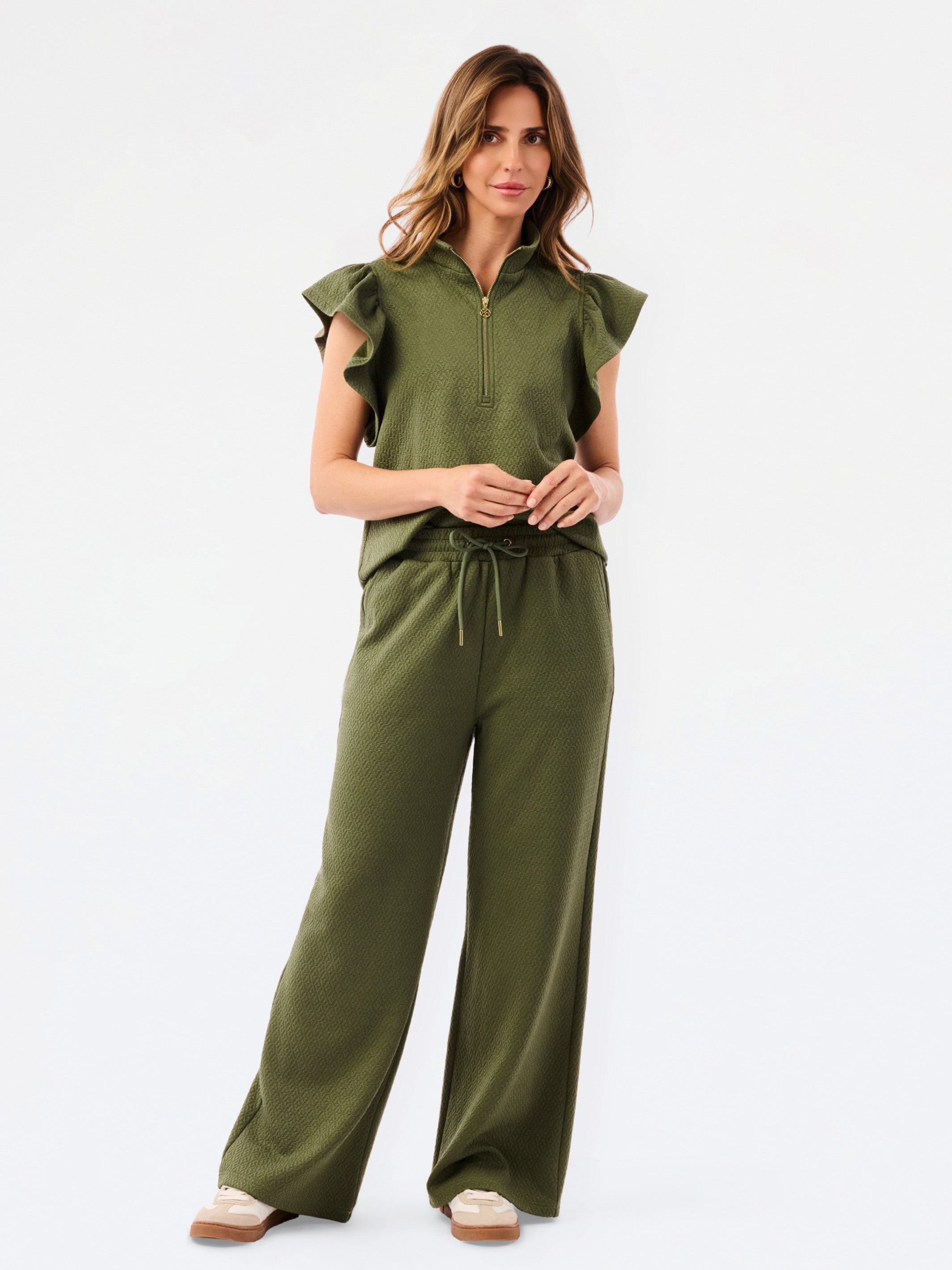 A woman modeling a matching olive green two-piece set consisting of a ruffled short-sleeve quarter-zip top and wide-leg pants, photographed against a clean white studio 