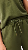 A woman modeling a matching olive green two-piece set consisting of a ruffled short-sleeve quarter-zip top and wide-leg pants, photographed against a clean white studio 