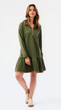 A woman modeling a short olive green textured dress with a half-zip neckline, ruffled hem, and gold trim, paired with sneakers against a plain white studio background.