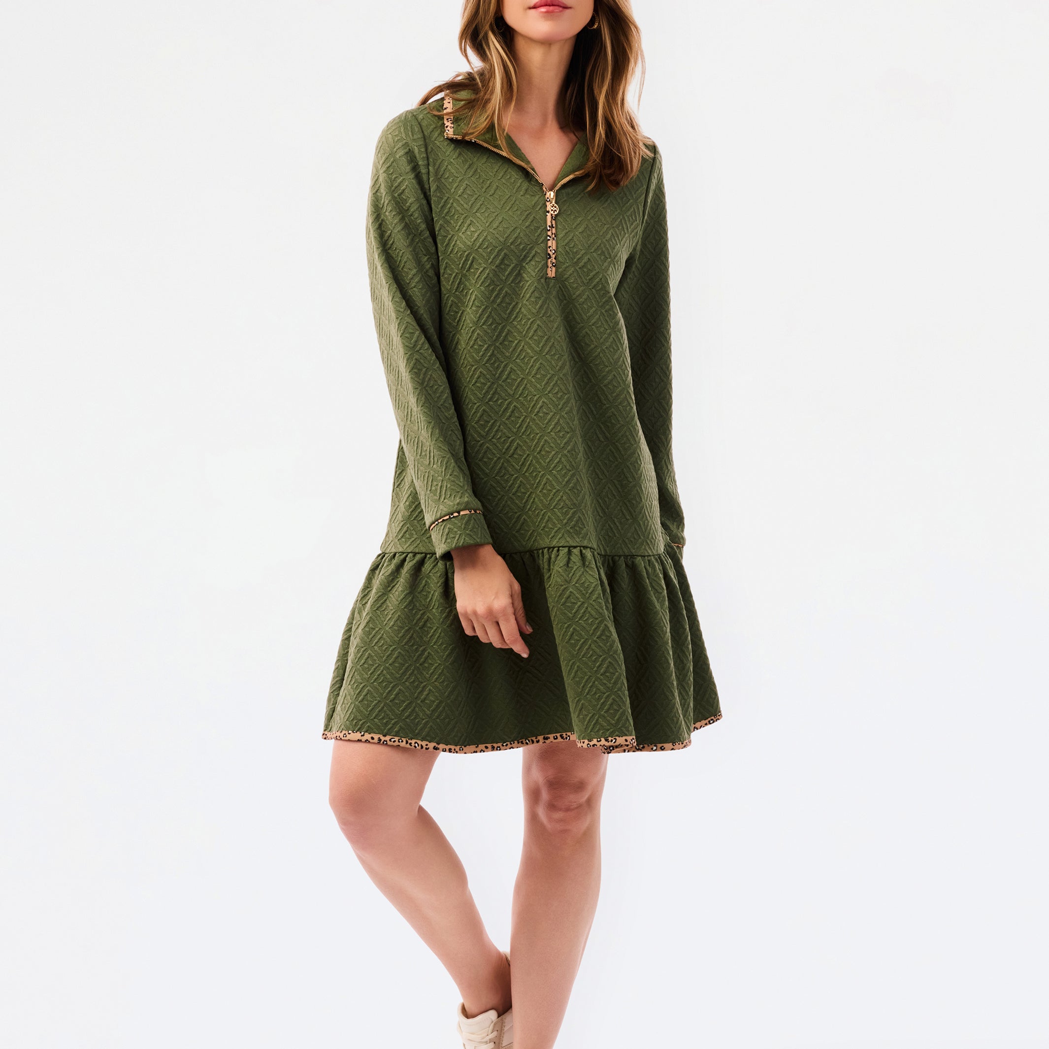 A woman modeling a short olive green textured dress with a half-zip neckline, ruffled hem, and gold trim, paired with sneakers against a plain white studio background.