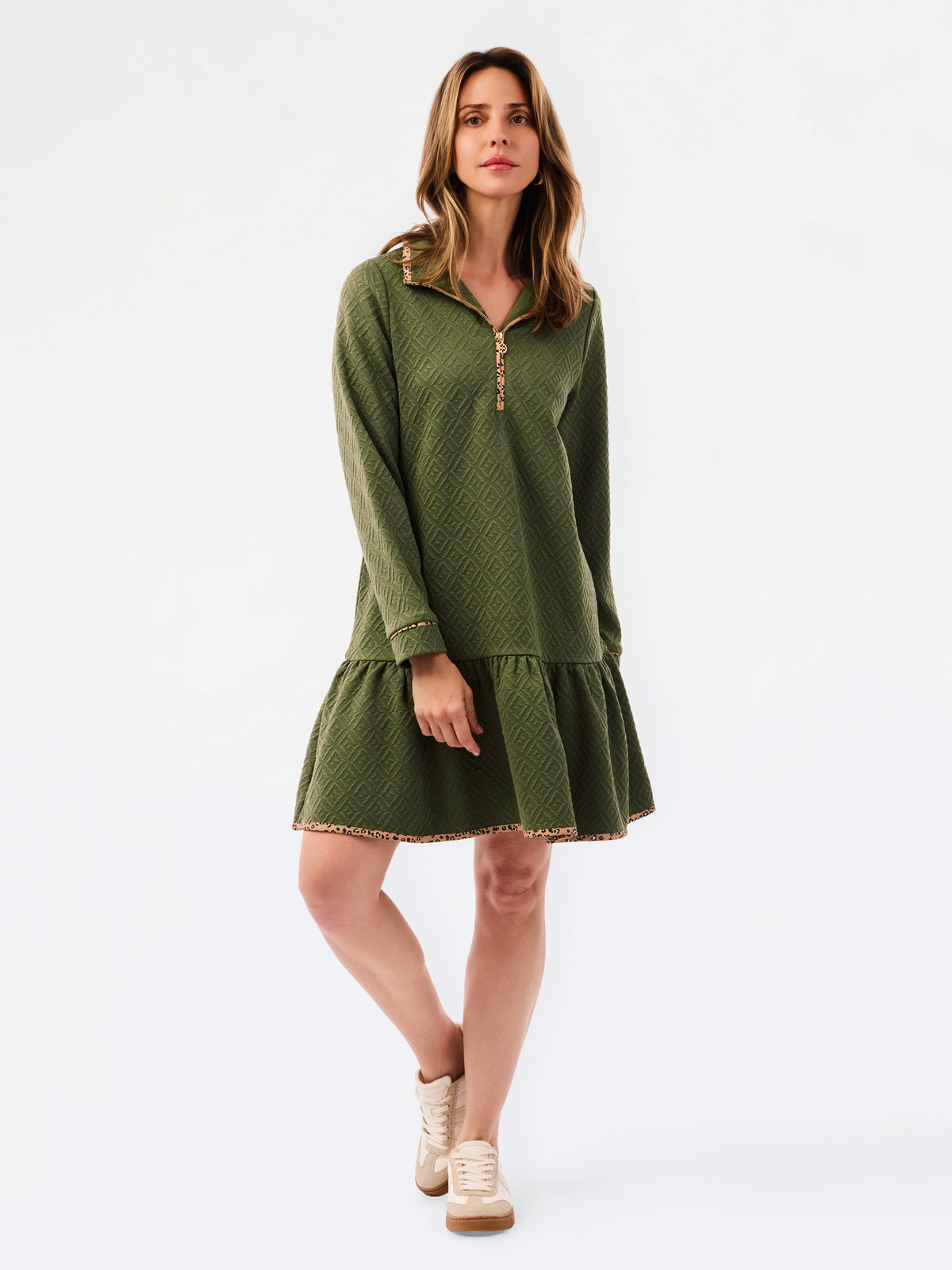 A woman modeling a short olive green textured dress with a half-zip neckline, ruffled hem, and gold trim, paired with sneakers against a plain white studio background.