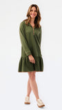 A woman modeling a short olive green textured dress with a half-zip neckline, ruffled hem, and gold trim, paired with sneakers against a plain white studio background.
