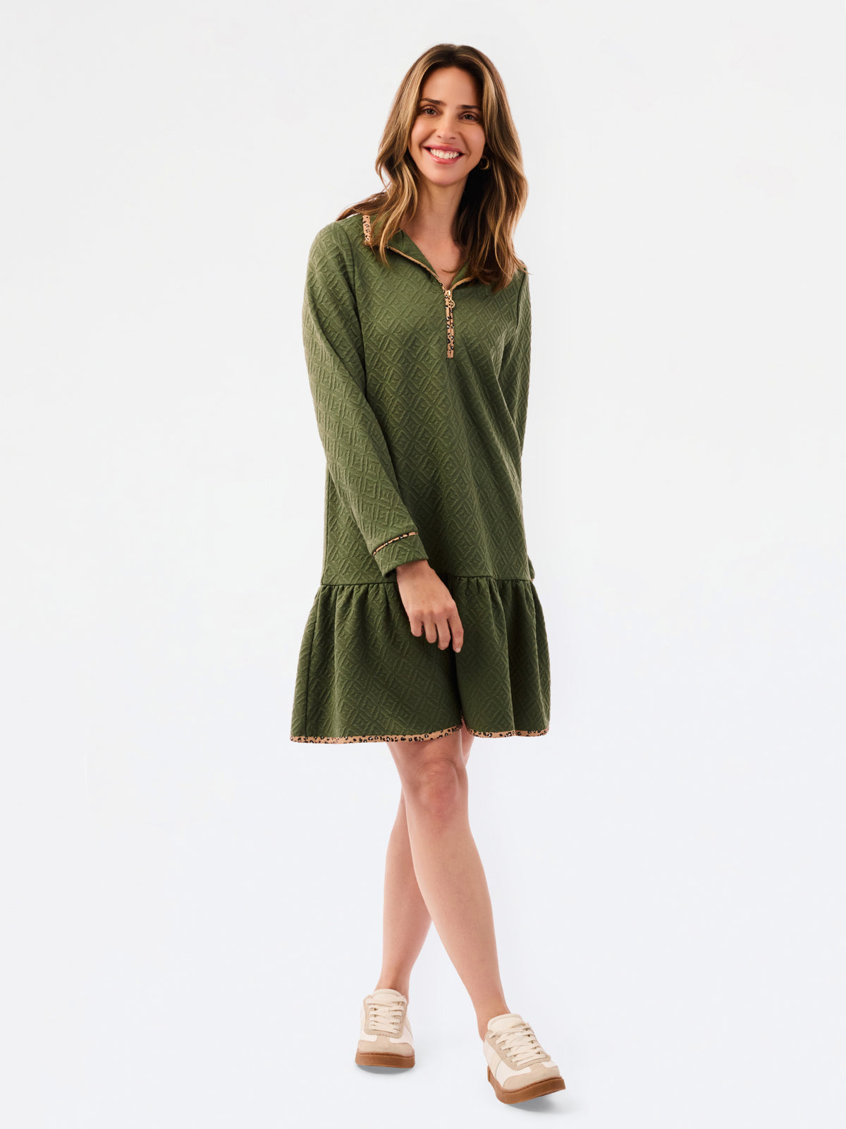 A woman modeling a short olive green textured dress with a half-zip neckline, ruffled hem, and gold trim, paired with sneakers against a plain white studio background.