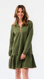 A woman modeling a short olive green textured dress with a half-zip neckline, ruffled hem, and gold trim, paired with sneakers against a plain white studio background.