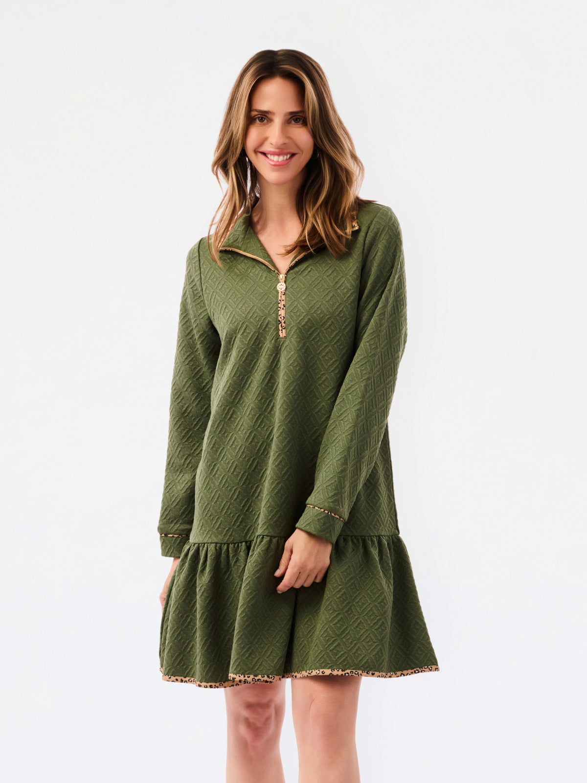 A woman modeling a short olive green textured dress with a half-zip neckline, ruffled hem, and gold trim, paired with sneakers against a plain white studio background.