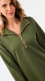 A woman modeling a short olive green textured dress with a half-zip neckline, ruffled hem, and gold trim, paired with sneakers against a plain white studio background.