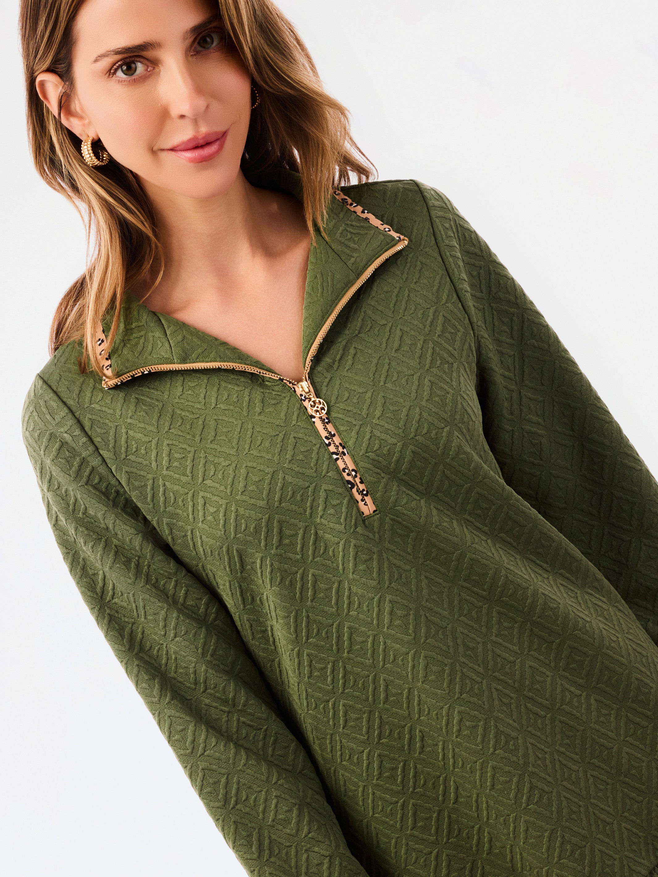 A woman modeling a short olive green textured dress with a half-zip neckline, ruffled hem, and gold trim, paired with sneakers against a plain white studio background.
