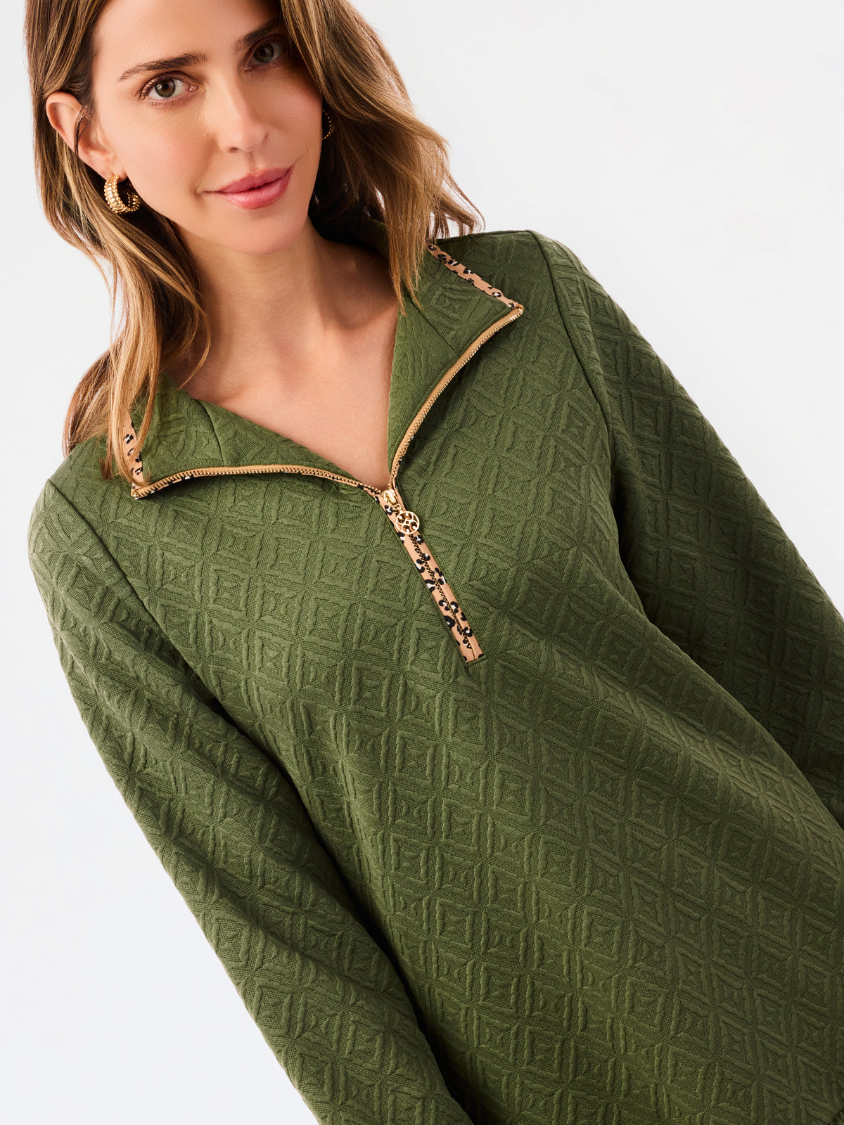 A woman modeling a short olive green textured dress with a half-zip neckline, ruffled hem, and gold trim, paired with sneakers against a plain white studio background.