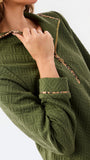 A woman modeling a short olive green textured dress with a half-zip neckline, ruffled hem, and gold trim, paired with sneakers against a plain white studio background.