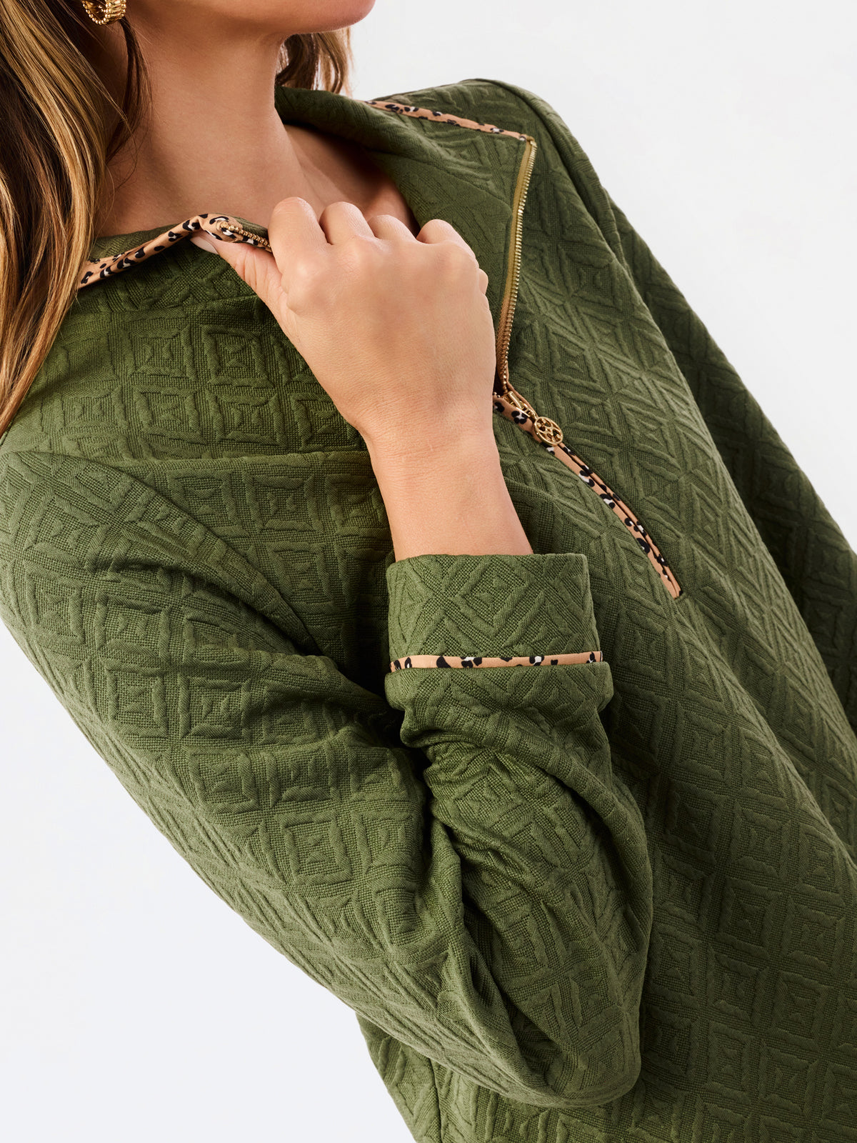 A woman modeling a short olive green textured dress with a half-zip neckline, ruffled hem, and gold trim, paired with sneakers against a plain white studio background.