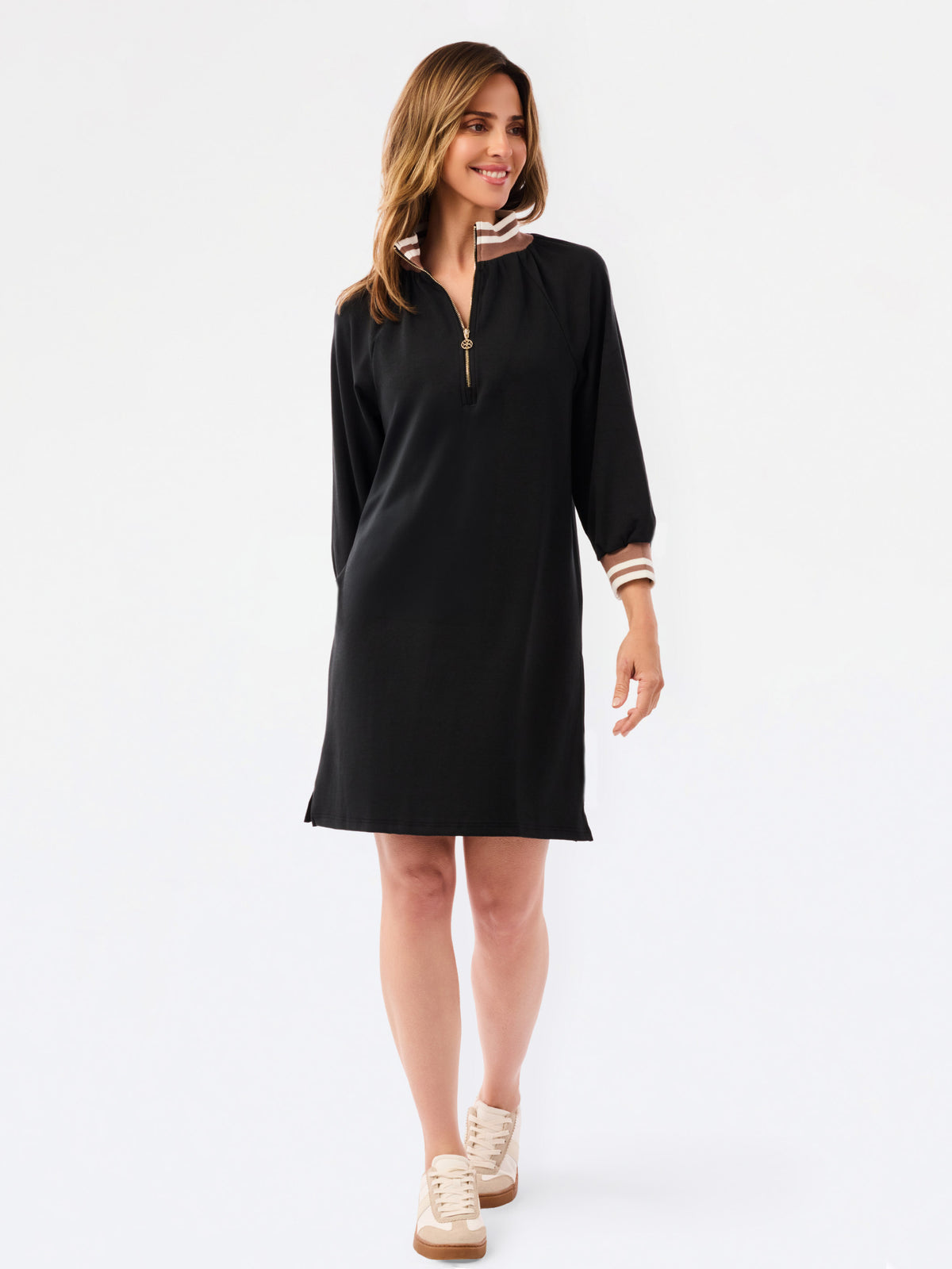 A woman modeling a black long-sleeve shift dress with a half-zip neckline and striped trim details on the collar and cuffs, set against a plain white studio background.