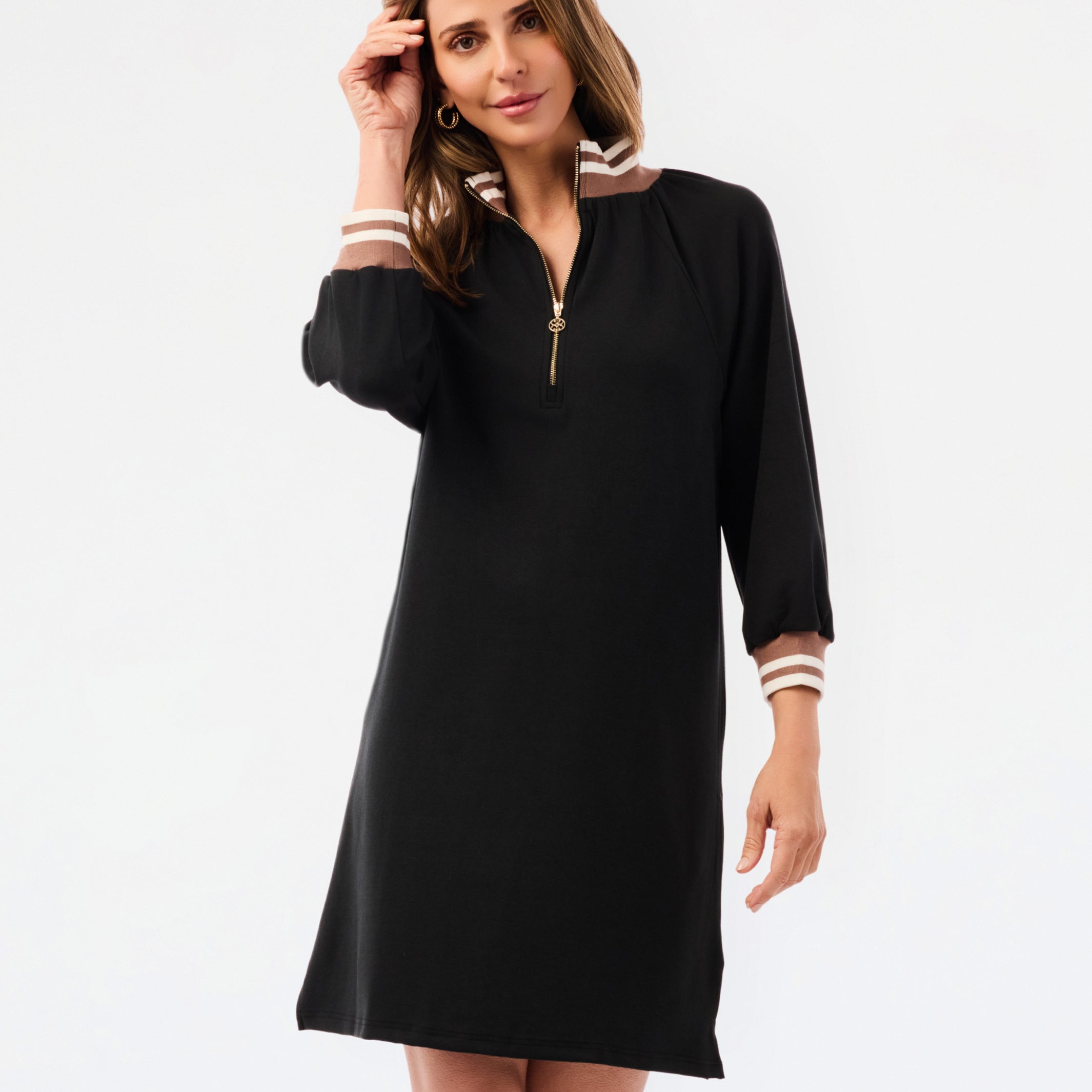 A woman modeling a black long-sleeve shift dress with a half-zip neckline and striped trim details on the collar and cuffs, set against a plain white studio background.