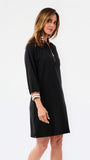 A woman modeling a black long-sleeve shift dress with a half-zip neckline and striped trim details on the collar and cuffs, set against a plain white studio background.
