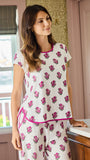 A woman wearing a two-piece short-sleeve pajama set with a white base, pink floral print, and pink trim, standing in a bedroom setting.