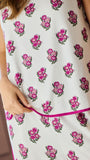 A close-up of a sleeveless white pajama set  with a repeating pink floral block print and a bright pink piped trim, worn by a model whose hand is visible adjusting the hem.