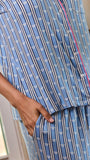 A close-up of a person wearing a blue pajama set with vertical white stripes and a small repeating motif, plus pink piping along the placket and waist. A hand is visible at the side pocket.