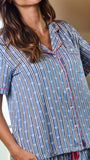 A woman wearing a short-sleeve pajama top with vertical blue, white, and black stripes and pink piping details, paired with matching striped pajama bottoms.