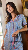 A woman wearing a blue and white striped short-sleeve pajama set with pink piping and a pink drawstring, standing in a bedroom next to a window and a wooden dresser.