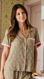A woman wearing a sage green short-sleeve pajama shirt with a pink floral pattern and pink trim, standing in a room with patterned wallpaper and a wall lamp.