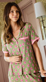 A woman wearing a sage green short-sleeve pajama set with a pink floral print and pink trim, standing in a softly lit bedroom.