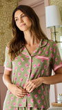 A woman wearing a sage green short-sleeve pajama shirt with a pink floral pattern and pink trim, standing in a room with patterned wallpaper and a wall lamp.