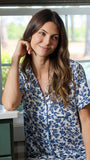 A woman wearing a short-sleeve button-front pajama set with a blue floral print on a white background, seated indoors on a teal sofa near a window.